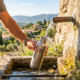 Randonneur remplissant une gourde en inox à une fontaine d'eau potable publique en pierre dans la campagne ensoleillée.